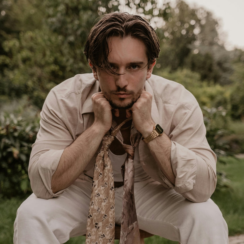 A man wearing a light beige button-up shirt and matching trousers, with a patterned beige tie loosely knotted around his neck, crouching outdoors in a garden setting.
