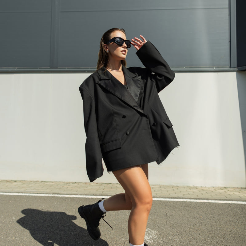 A woman wearing an oversized black double-breasted blazer, matching black mini skirt, black sunglasses, and black boots walks outdoors against a gray and white wall.