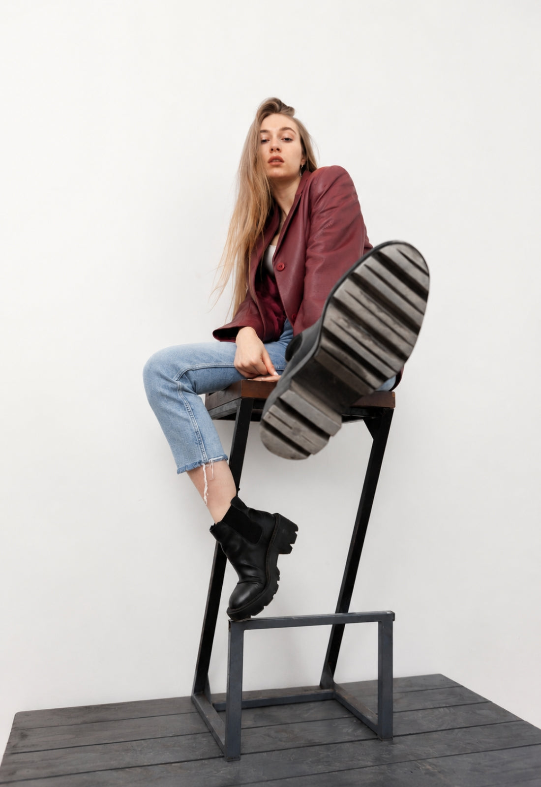 A woman sits on a black metal stool against a white wall, wearing a burgundy blazer, light blue jeans, and black ankle boots. She is posed with one leg extended upward, showing the striped pattern of the stool's seat cushion.