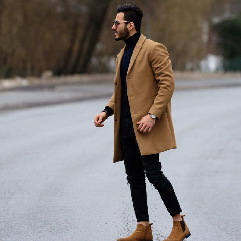 A man walking outdoors wearing a camel-colored overcoat, black turtleneck, black pants, and tan ankle boots.