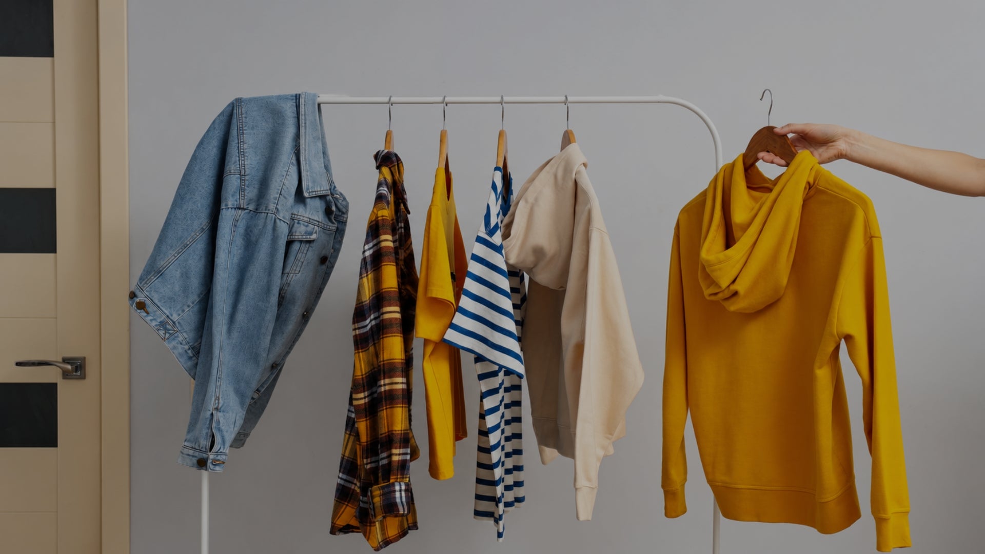 A clothing rack displaying a variety of apparel items, including a blue denim jacket, a yellow plaid shirt, a yellow top, a beige cardigan, a striped shirt, and a mustard yellow hoodie being held by a hand on the right.