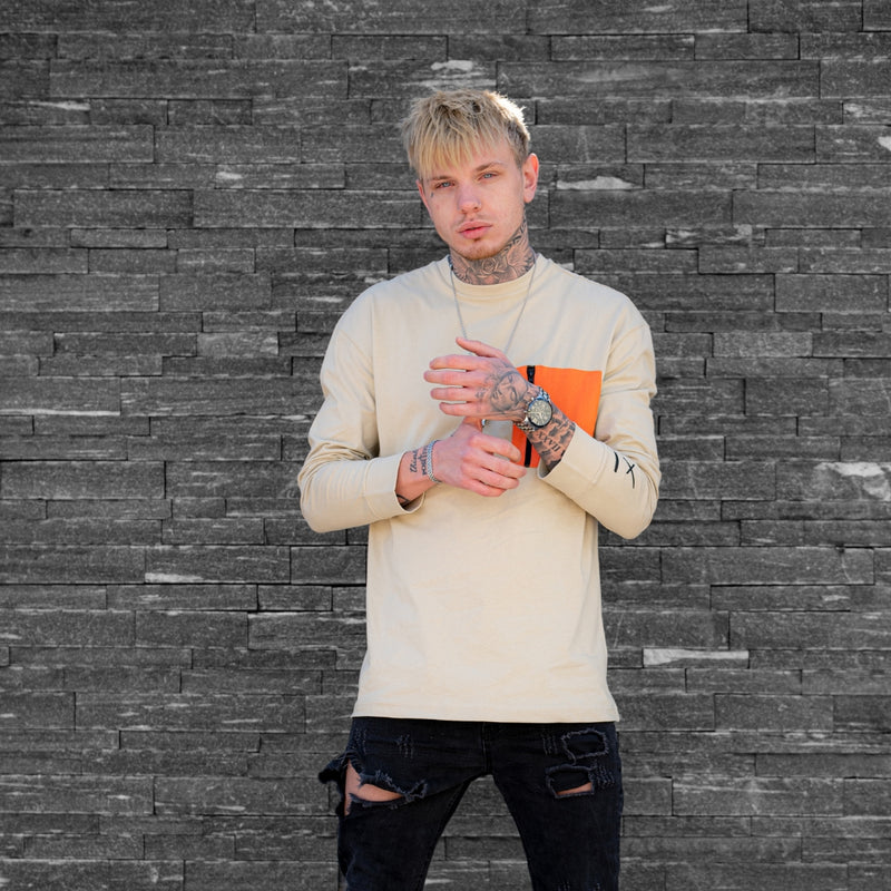 A man with blonde hair and tattoos stands against a gray stone wall, wearing a cream-colored long-sleeve shirt with a bright orange rectangular accent on the chest, paired with dark ripped jeans.