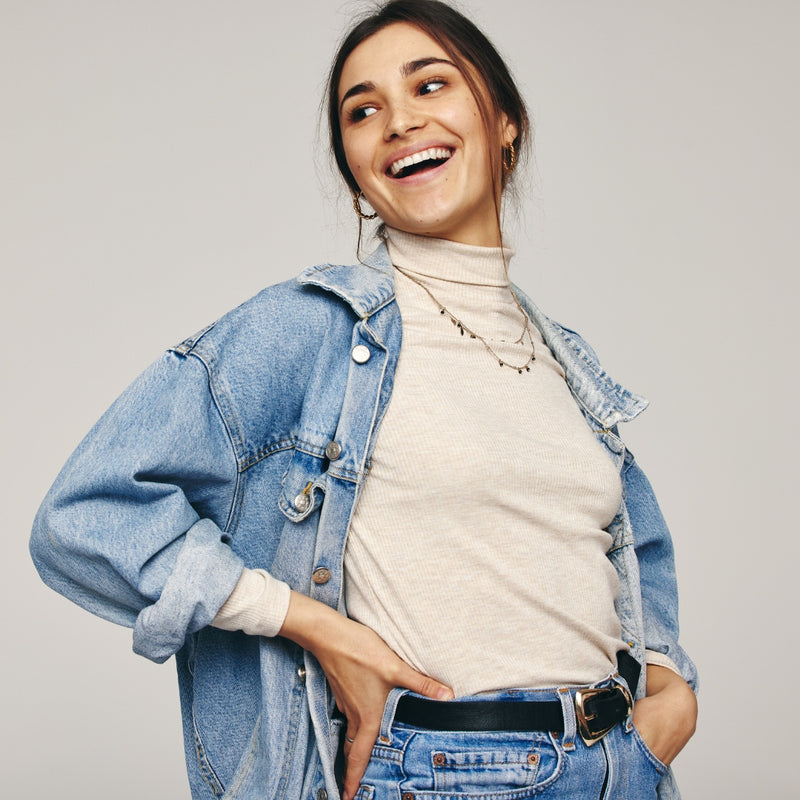 A woman smiling and looking to the side, wearing a light blue denim jacket over a cream turtleneck, paired with blue jeans and a black belt, against a solid light gray background.