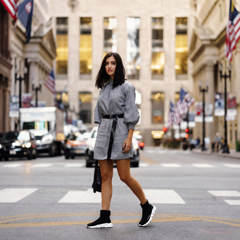 A woman walks across a city street wearing a gray mini shirt dress with a black belt and black ankle socks with white sneakers.