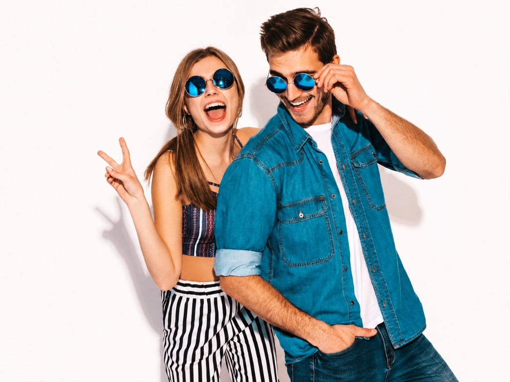 A man and a woman posing together against a white background, both wearing blue mirrored sunglasses, a blue denim shirt, and striped pants.