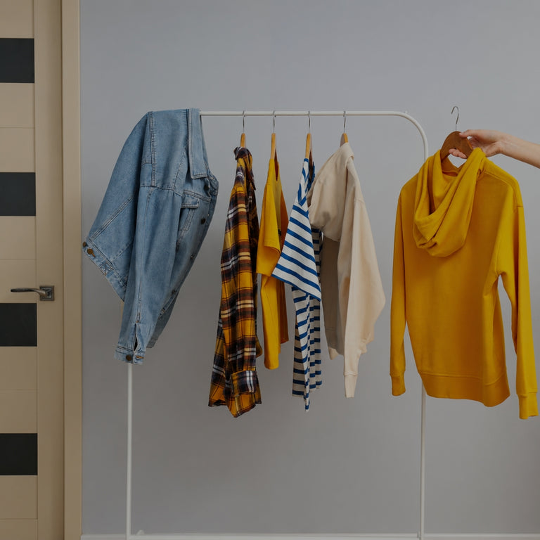 A clothing rack displaying a variety of apparel items, including a blue denim jacket, a yellow plaid shirt, a yellow top, a beige cardigan, a striped shirt, and a mustard yellow hoodie being held by a hand on the right.