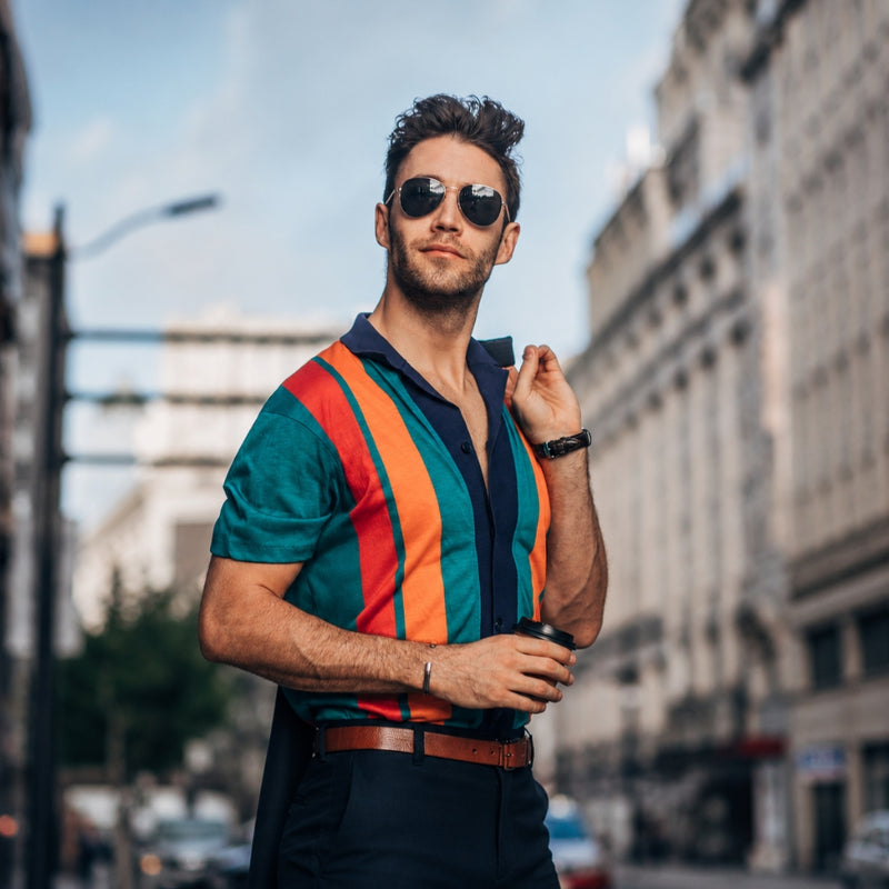 A man wearing a colorful striped short-sleeve polo shirt with teal, orange, and red vertical stripes, styled with dark trousers, sunglasses, and a watch, standing on a city street.