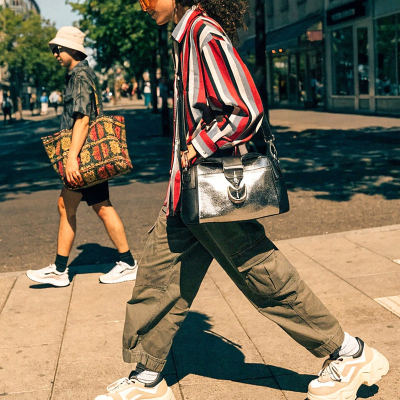 A person walking on a sunny city sidewalk wearing a red, white, and black striped button-up shirt, olive cargo pants, white sneakers, and carrying a shiny black shoulder bag. Another person is visible in the background carrying a patterned tote bag.