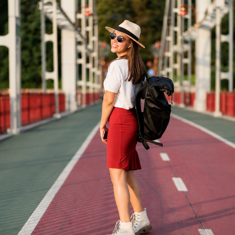 A woman stands on a red and green bridge wearing a white t-shirt, a red knee-length skirt, a beige fedora hat, sunglasses, and carrying a black backpack.